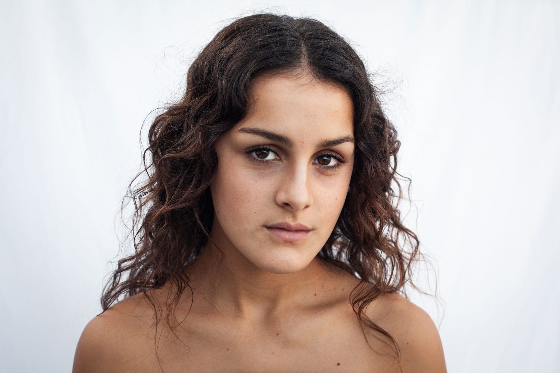 Una dona jove amb els cabells castanys ondulats i maquillatge natural mira directament a la c&agrave;mera sobre un fons blanc llis. T&eacute; una expressi&oacute; neutra i les espatlles nues.