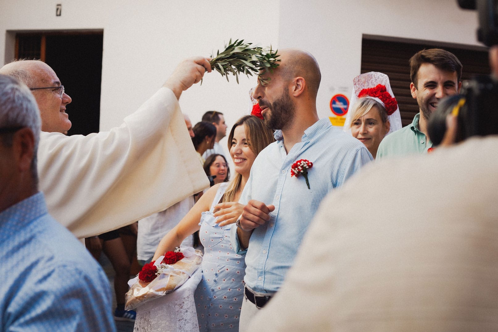 Un sacerdot beneeix un home somrient amb una branca d'olivera durant una cerim&ograve;nia a l'aire lliure, envoltat de gent, alguns dels quals sostenen flors i porten roba festiva.