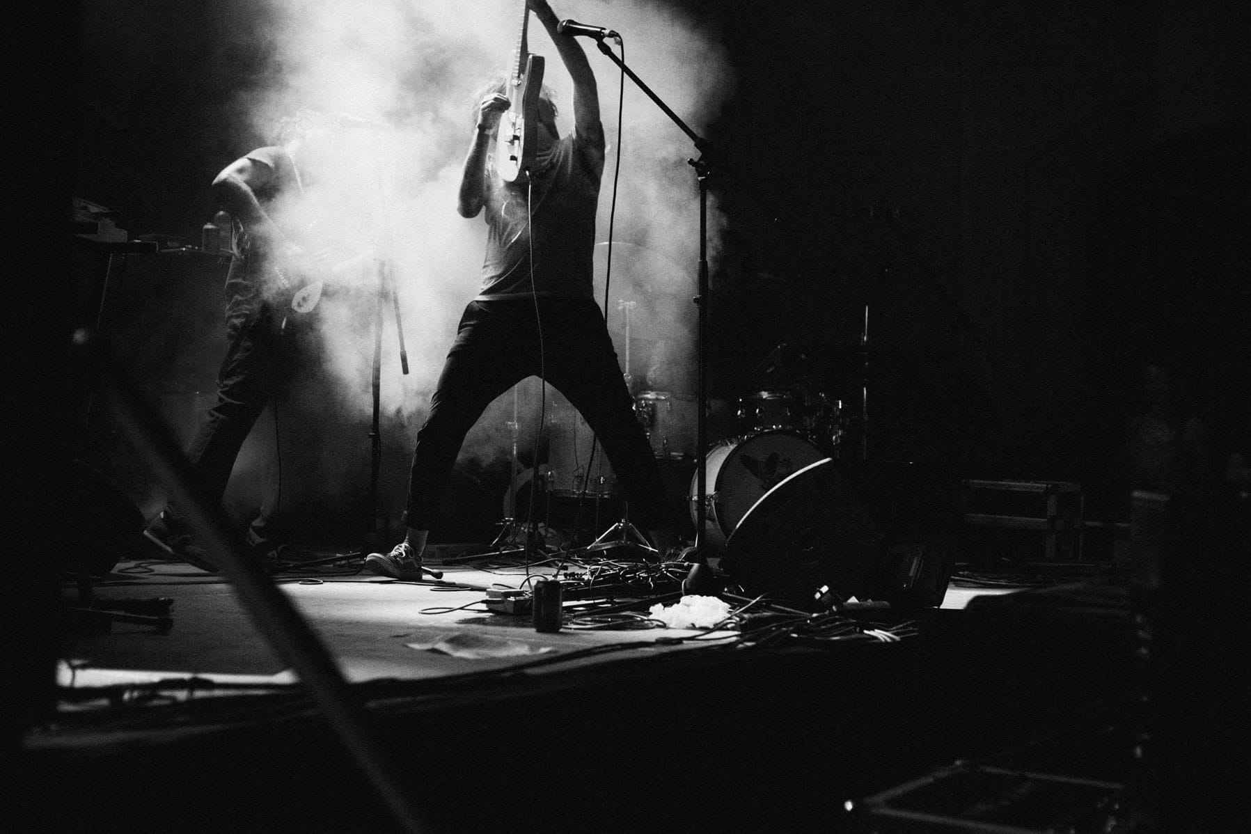 Un m&uacute;sic a l'escenari toca apassionadament una guitarra el&egrave;ctrica entre un fum dram&agrave;tic i llums de l'escenari, amb una bateria i un altre int&egrave;rpret visibles al fons. L'atmosfera &eacute;s en&egrave;rgica i intensa.