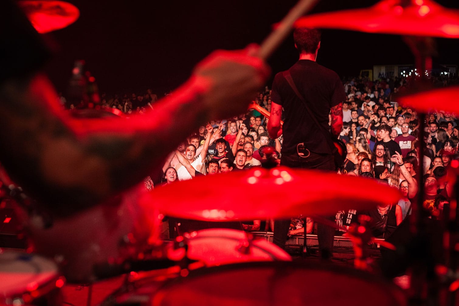 El bra&ccedil; i la bateria d'un bateria estan en primer pla, desenfocats, amb un guitarrista dret davant d'una multitud que anima sota els llums vermells de l'escenari en un concert en directe.