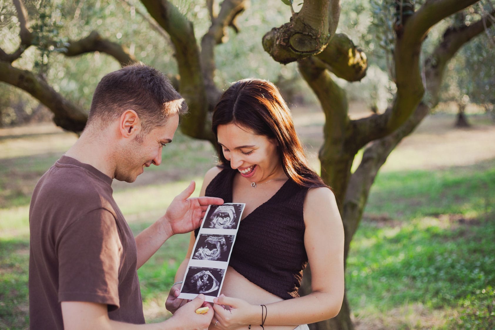 Una parella somrient es troba a l'aire lliure al costat d'un arbre, mirant feli&ccedil;ment les imatges de l'ecografia, amb la dona tocant-li suaument la panxa, cosa que suggereix que esperen un nad&oacute;.