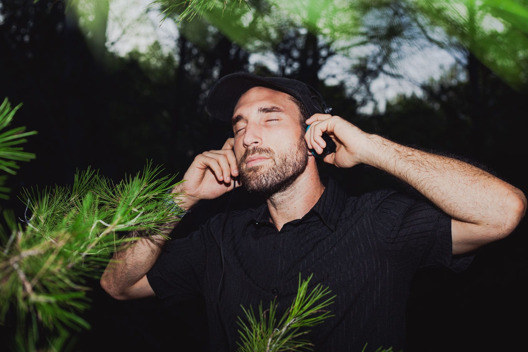 Un home amb barba i gorra es troba entre branques verdes de pi, amb els ulls tancats i somrient lleugerament, escoltant amb auriculars i semblant gaudir de la m&uacute;sica en un entorn forestal.